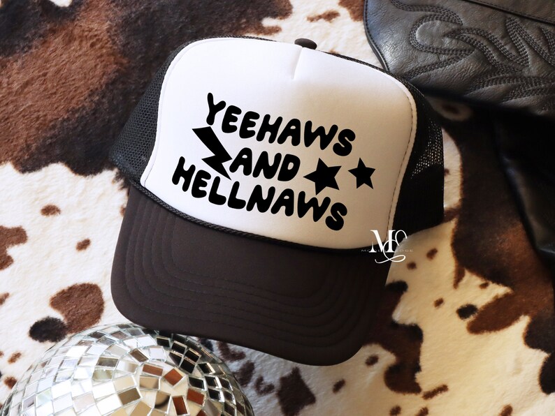May include: White trucker hat with a black mesh back. The front of the hat has black text that reads "Yeehaws and Hellnaws" with a lightning bolt and two stars.