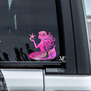 May include: Pink mermaid sticker with a pink starfish on a black car window. The mermaid is sitting with her tail curled around her.