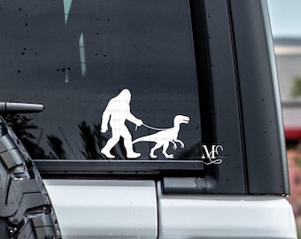 May include: White vinyl decal featuring a Bigfoot figure walking a dinosaur on a leash. The decal is applied to a vehicle window, with a tire visible in the lower left corner. The design includes the initials "MC" in a cursive font.