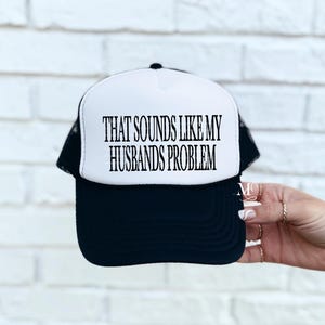 May include: A black and white trucker hat with the text "THAT SOUNDS LIKE MY HUSBANDS PROBLEM" in black. The hat has a white front panel and a black brim and mesh back.