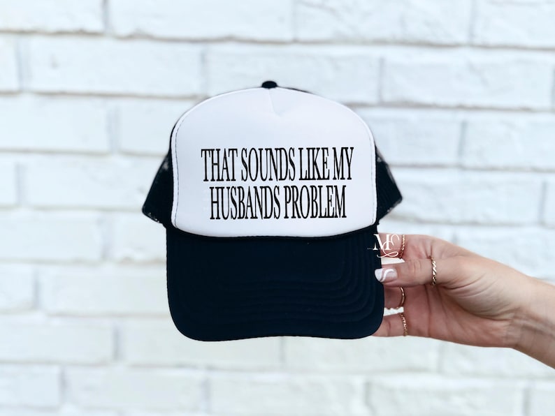 May include: A black and white trucker hat with the text "THAT SOUNDS LIKE MY HUSBANDS PROBLEM" in black. The hat has a white front panel and a black brim and mesh back.