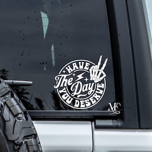 May include: White vinyl decal with the text "Have The Day You Deserve" in a circular design. The design includes a skeleton hand making a peace sign, a lightning bolt, and stars. The decal is affixed to a vehicle window.