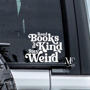 May include: White vinyl decal with the words "Read Books, Be Kind, Stay Weird" in a decorative font. The decal is affixed to the rear window of a vehicle, with a tire visible in the foreground.