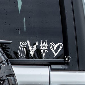 May include: White vinyl decal featuring a salt shaker, sprigs, lavender, and a heart. The decal is applied to a dark vehicle window. The design is minimalist and likely for car decoration.
