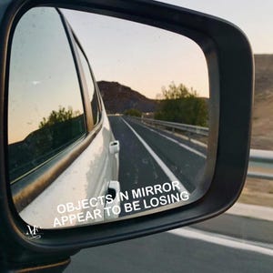 May include: A car's wing mirror showing a road with a white car, a highway, and a mountain range in the distance. The text "OBJECTS IN MIRROR APPEAR TO BE LOSING" is visible on the mirror.