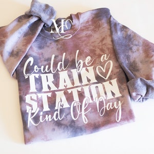 May include: A purple and gray tie-dye sweatshirt with the text "Could be a train station kind of day" in white lettering with a white heart.