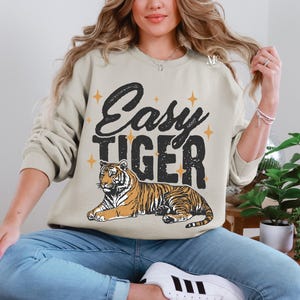 K&ouml;nnte beinhalten: Ein beiger Pullover mit dem Text "Easy Tiger" in schwarzen Buchstaben und einer stilisierten Tigerillustration. Der Tiger ist orange und schwarz mit gelbem Hintergrund.