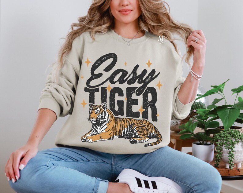 K&ouml;nnte beinhalten: Ein beiger Pullover mit dem Text "Easy Tiger" in schwarzen Buchstaben und einer stilisierten Tigerillustration. Der Tiger ist orange und schwarz mit gelbem Hintergrund.