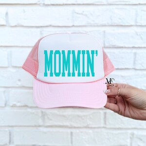 Puede incluir: Gorra de camionero rosa y blanca con un gr&aacute;fico turquesa 'MOMMIN'.