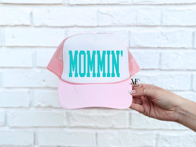 Puede incluir: Gorra de camionero rosa y blanca con un gr&aacute;fico turquesa 'MOMMIN'.