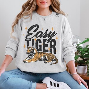 K&ouml;nnte beinhalten: Ein grauer Pullover mit dem Text "Easy Tiger" in schwarzen Buchstaben und einer stilisierten Tigerillustration. Der Tiger ist orange und schwarz auf wei&szlig;em Hintergrund.