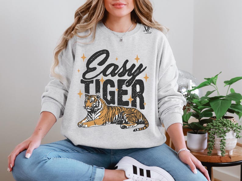 K&ouml;nnte beinhalten: Ein grauer Pullover mit dem Text "Easy Tiger" in schwarzen Buchstaben und einer stilisierten Tigerillustration. Der Tiger ist orange und schwarz auf wei&szlig;em Hintergrund.