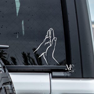 May include: White vinyl decal of a hand giving a paw a high five, applied to a vehicle window. The design features a dog paw and a human hand, with the initials "MC" in a script font. The image is set against a dark background.