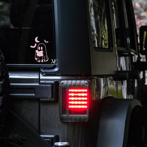 May include: A pink ghost and moon decal on a black vehicle window. The decal features a ghost with a friendly face, two crescent moons, and the initials "MC". The vehicle has a red taillight.