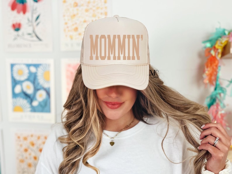 Puede incluir: Una gorra de camionero beige con una malla trasera marr&oacute;n. La gorra tiene la palabra "MOMMIN'" bordada en la parte delantera en una fuente marr&oacute;n.