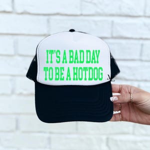 May include: A white and black trucker hat with a green text graphic that reads "It's a bad day to be a hotdog".