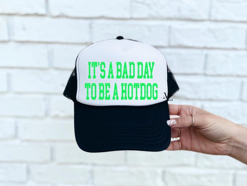 May include: A white and black trucker hat with a green text graphic that reads "It's a bad day to be a hotdog".