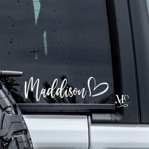 May include: A white vinyl decal applied to a dark vehicle window, featuring the name Maddison in a cursive script font followed by a hand-drawn heart outline. A small logo with the letters MC is visible in the lower right corner of the glass.