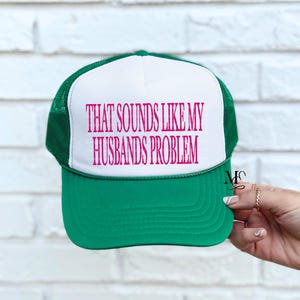 May include: A green and white trucker hat with the text "THAT SOUNDS LIKE MY HUSBANDS PROBLEM" in pink. The hat has a green brim and mesh back, with a white front panel. The text is centered on the front.