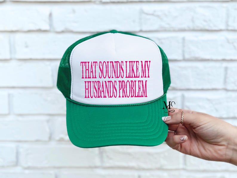 May include: A green and white trucker hat with the text "THAT SOUNDS LIKE MY HUSBANDS PROBLEM" in pink. The hat has a green brim and mesh back, with a white front panel. The text is centered on the front.