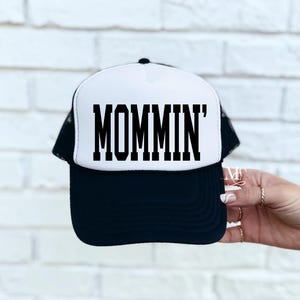 Puede incluir: Una gorra de camionero negra y blanca con un panel frontal blanco y una malla negra en la parte posterior. El panel frontal tiene el texto "MOMMIN'" en letras negras en bloque.