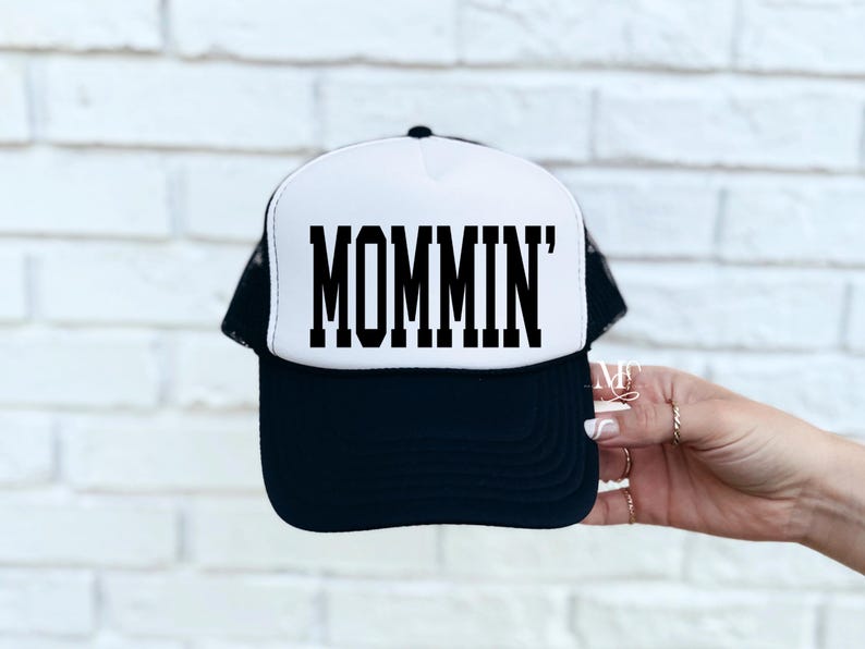 Puede incluir: Una gorra de camionero negra y blanca con un panel frontal blanco y una malla negra en la parte posterior. El panel frontal tiene el texto "MOMMIN'" en letras negras en bloque.