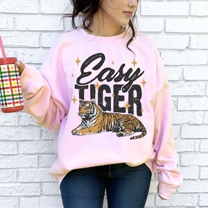 K&ouml;nnte beinhalten: Ein rosa Sweatshirt mit dem Text "Easy Tiger" in schwarzen Buchstaben und einer Cartoon-Tiger-Illustration. Der Tiger ist orange und schwarz mit wei&szlig;em Hintergrund.
