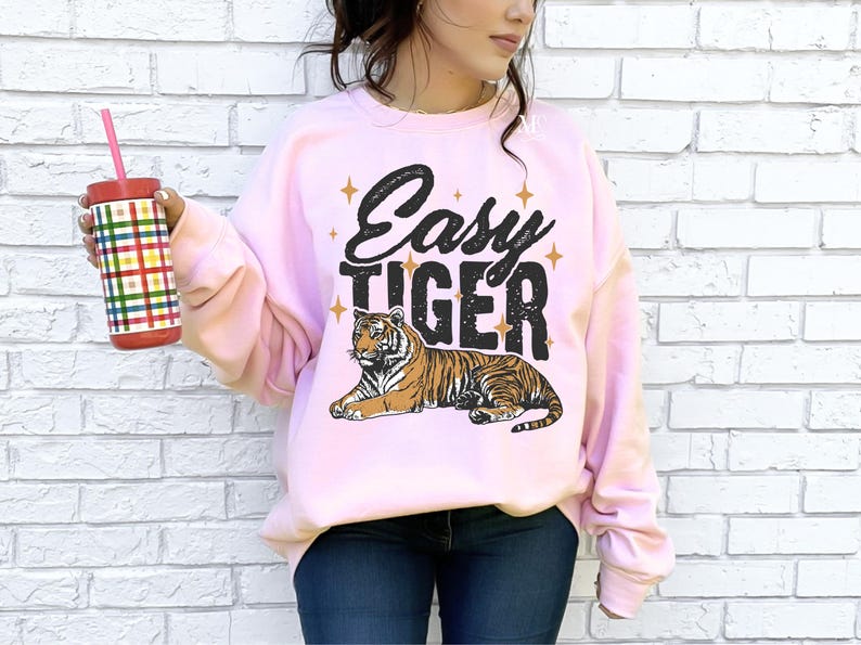 K&ouml;nnte beinhalten: Ein rosa Sweatshirt mit dem Text "Easy Tiger" in schwarzen Buchstaben und einer Cartoon-Tiger-Illustration. Der Tiger ist orange und schwarz mit wei&szlig;em Hintergrund.