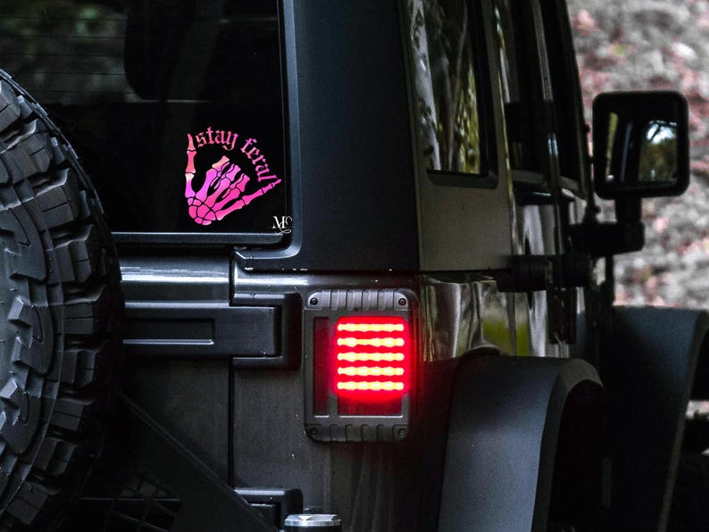 May include: A black Jeep with a pink and white "stay feral" sticker on the back window. The Jeep has a spare tire on the back and a red taillight.