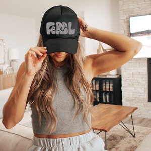 以下が含まれることがあります： レトロなフォントで白い「FERAL」ロゴが入った黒いトラッカーハット。長い茶色の髪をした人が被っています。