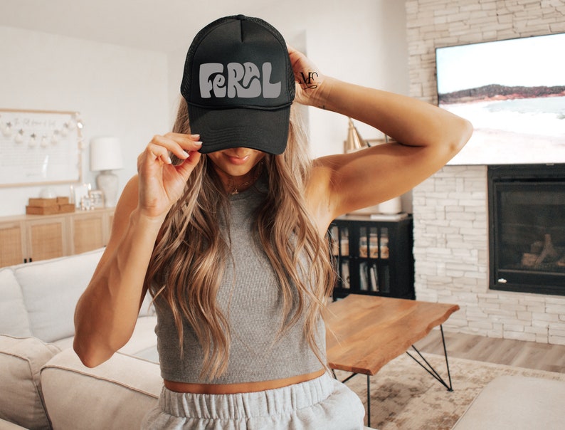以下が含まれることがあります： レトロなフォントで白い「FERAL」ロゴが入った黒いトラッカーハット。長い茶色の髪をした人が被っています。