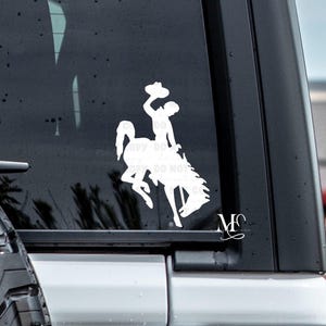 May include: White vinyl decal of a rodeo cowboy riding a bucking bronco, with the cowboy holding his hat up in the air. The decal is applied to a vehicle window. The image includes the initials "MC" in a cursive font.