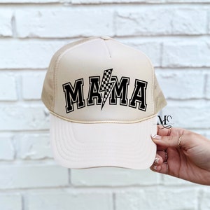 May include: A beige trucker hat with a black and white checkered lightning bolt and the word "MAMA" in black block letters.