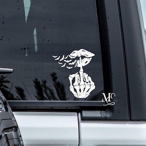 May include: White vinyl decal featuring a skeleton hand giving the middle finger, with lips and bats above. The decal is applied to a black vehicle window. A stylized "MC" logo is in the lower right corner.