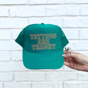 May include: A green trucker hat with a white mesh back. The hat has the text "Tattoos Are Trashy" in orange lettering.