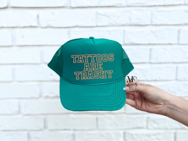May include: A green trucker hat with a white mesh back. The hat has the text "Tattoos Are Trashy" in orange lettering.
