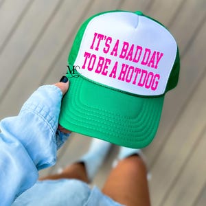 May include: A green and white trucker hat with the text "It's a bad day to be a hotdog" in pink lettering.