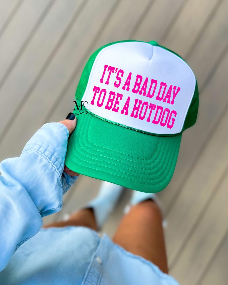 May include: A green and white trucker hat with the text "It's a bad day to be a hotdog" in pink lettering.