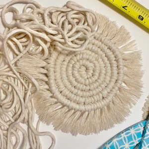 May include: A close-up of a white macrame coaster with a fringe border. The coaster is made with a thick, natural fibre cord and has a circular design. There is also a ball of cord and a pair of scissors in the image.