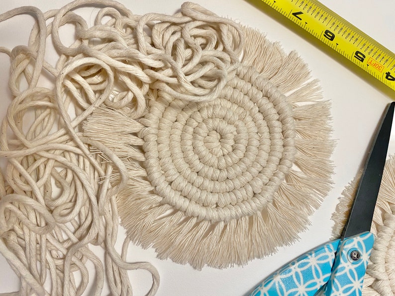 Pode incluir: Um close-up de um descanso de copo de macram&eacute; branco com uma borda de franja. O descanso de copo &eacute; feito com um cord&atilde;o grosso de fibra natural e tem um design circular. Tamb&eacute;m h&aacute; uma bola de cord&atilde;o e um par de tesouras na imagem.
