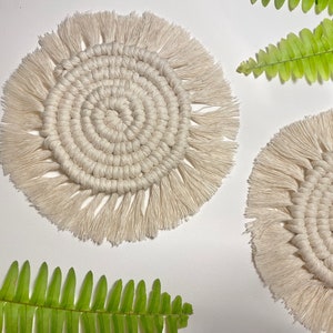 Pode incluir: Dois  copos de croch&ecirc; branco com detalhes em franjas. Os copos s&atilde;o redondos e t&ecirc;m um padr&atilde;o circular no centro. Os copos est&atilde;o sobre uma superf&iacute;cie branca com folhas de samambaia verdes.