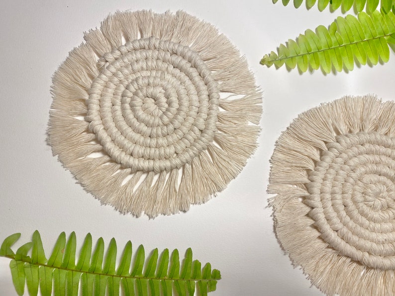 Pode incluir: Dois  copos de croch&ecirc; branco com detalhes em franjas. Os copos s&atilde;o redondos e t&ecirc;m um padr&atilde;o circular no centro. Os copos est&atilde;o sobre uma superf&iacute;cie branca com folhas de samambaia verdes.