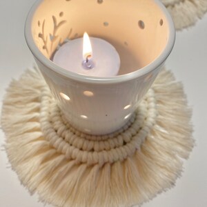 Pode incluir: Um porta-velas de cer&acirc;mica branca com um design decorativo e uma vela acesa dentro, sentado em um descanso de copo de croch&ecirc; de cor creme com franjas.