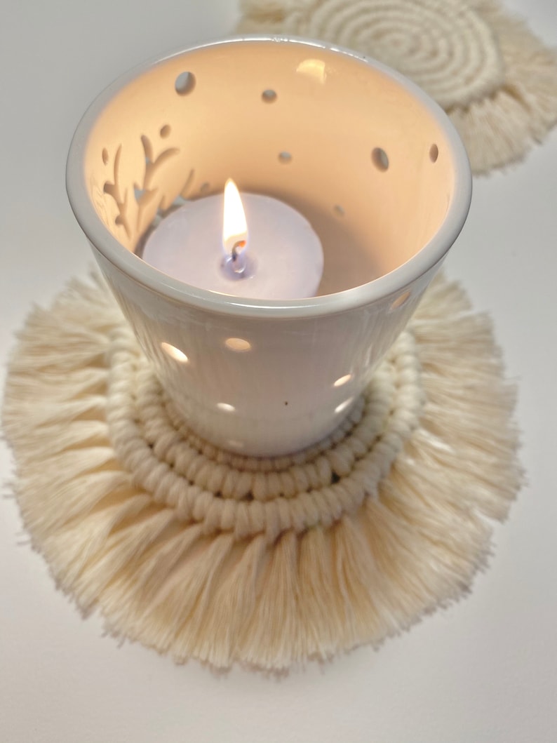 Pode incluir: Um porta-velas de cer&acirc;mica branca com um design decorativo e uma vela acesa dentro, sentado em um descanso de copo de croch&ecirc; de cor creme com franjas.