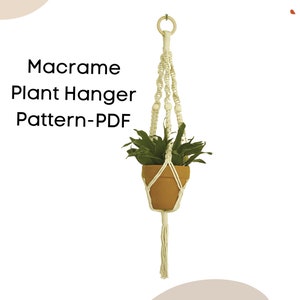 May include: A macrame plant hanger with a terracotta pot holding a green plant. The off-white macrame design includes a wooden ring at the top and a tassel at the bottom. Text on the image reads "Macrame Plant Hanger Pattern-PDF".