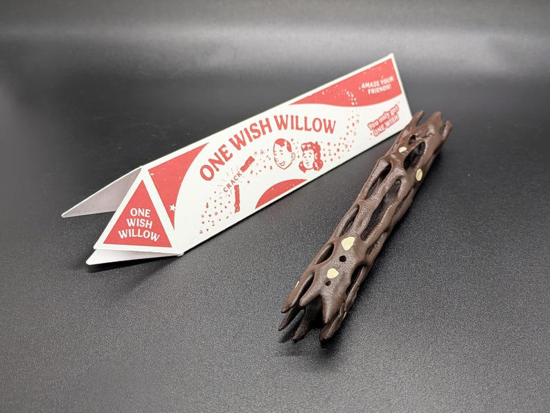 May include: A dark brown, textured, cylindrical object with an open lattice structure and small light-colored circular inlays, accompanied by a triangular white and red paper packaging labeled ONE WISH WILLOW with illustrated faces.