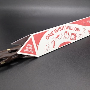 May include: A brown, textured, twig-like object partially inside a triangular white and red cardboard box. The packaging features the text "ONE WISH WILLOW," "AMAZE YOUR FRIENDS!," and "You only get ONE WISH," along with illustrations of two faces.