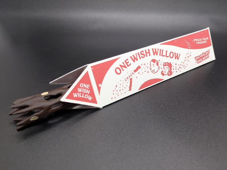 May include: A brown, textured, twig-like object partially inside a triangular white and red cardboard box. The packaging features the text "ONE WISH WILLOW," "AMAZE YOUR FRIENDS!," and "You only get ONE WISH," along with illustrations of two faces.