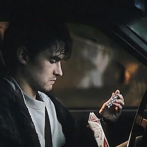 May include: A person with dark hair wearing a light gray crew neck shirt and a dark textured jacket sits in the driver seat of a vehicle, holding a small silver metallic object in one hand and a white and red rectangular package in the other.