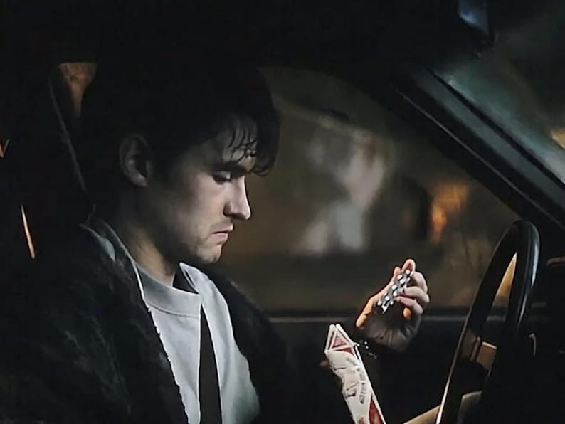May include: A person with dark hair wearing a light gray crew neck shirt and a dark textured jacket sits in the driver seat of a vehicle, holding a small silver metallic object in one hand and a white and red rectangular package in the other.